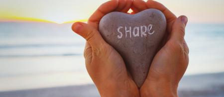 Two hands holding a heart shaped stone that says Share with a lake in the background