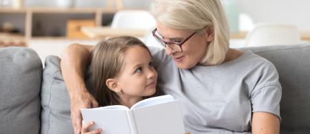 Senior woman sitting on a sofa with her arm around a little girl reading a story to her.