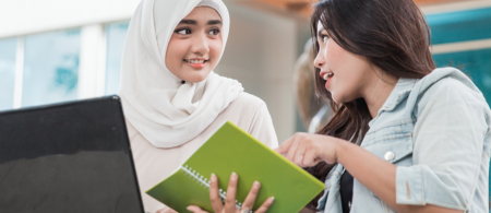 Muslim and American girls looking at a book and computer together.