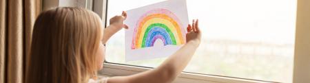 Child holding a drawing of a rainbow up against a window