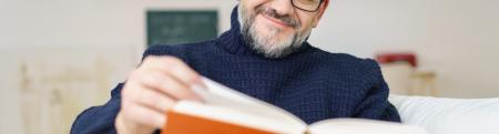 Man with glasses and a beard reading a book with an orange cover.