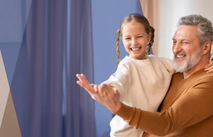 Man holds his granddaughter with arms extended while they laugh in their home