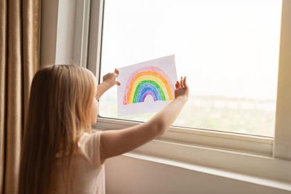 Child holding a drawing of a rainbow up against a window